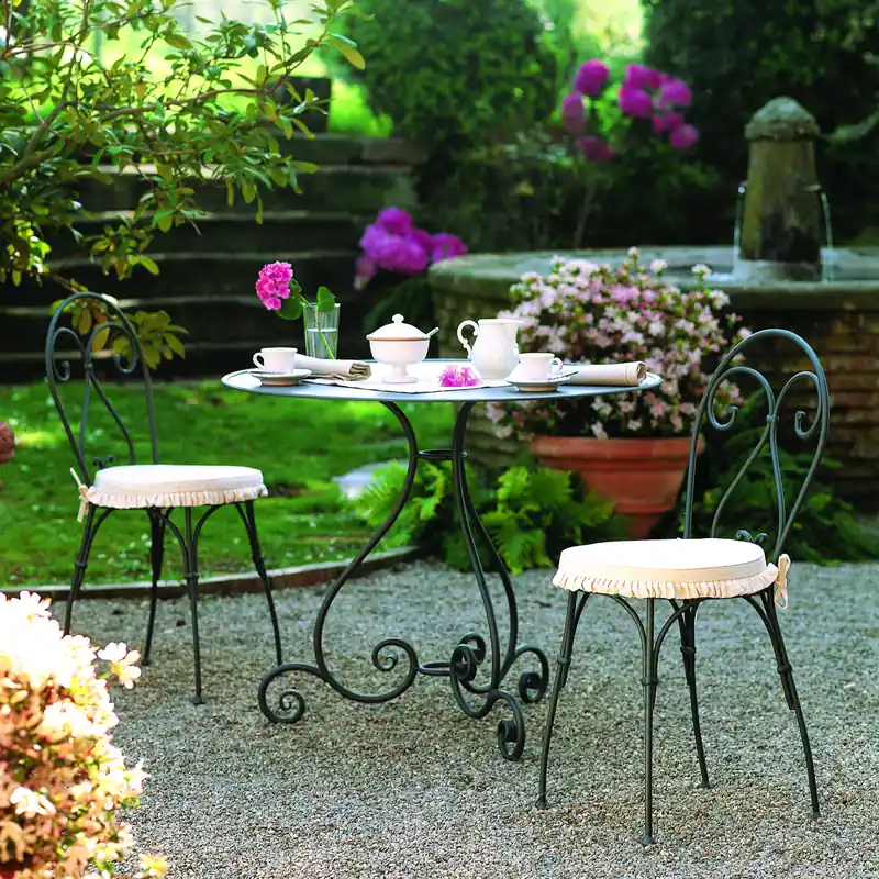 Unopiù Aurora wrought-iron round table set for tea with two stools on gravel, in a garden with flowering pots