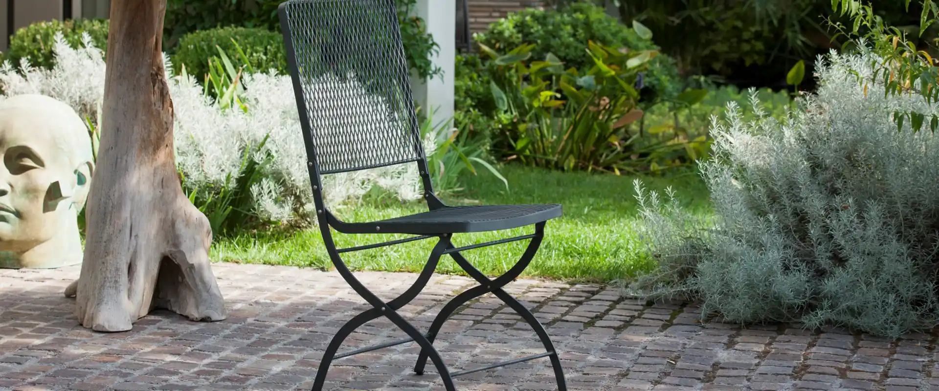 Unopiù Aurora folding iron chair beside a stone table on a cobbled garden path, with a lawn and shrubs