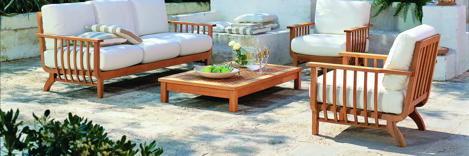 Unopiù Chelsea teak sofa, armchairs and coffee table with white cushions on a sunlit terrace with whitewashed walls and lush plants