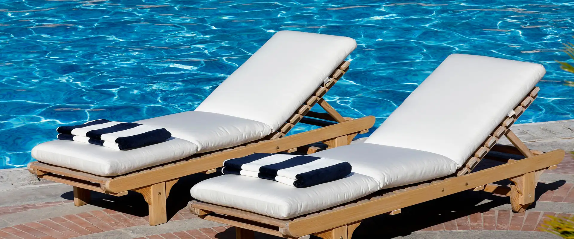 Two Unopiù Chelsea teak sun loungers with white mattresses and rolled towels on a wooden deck beside the sea