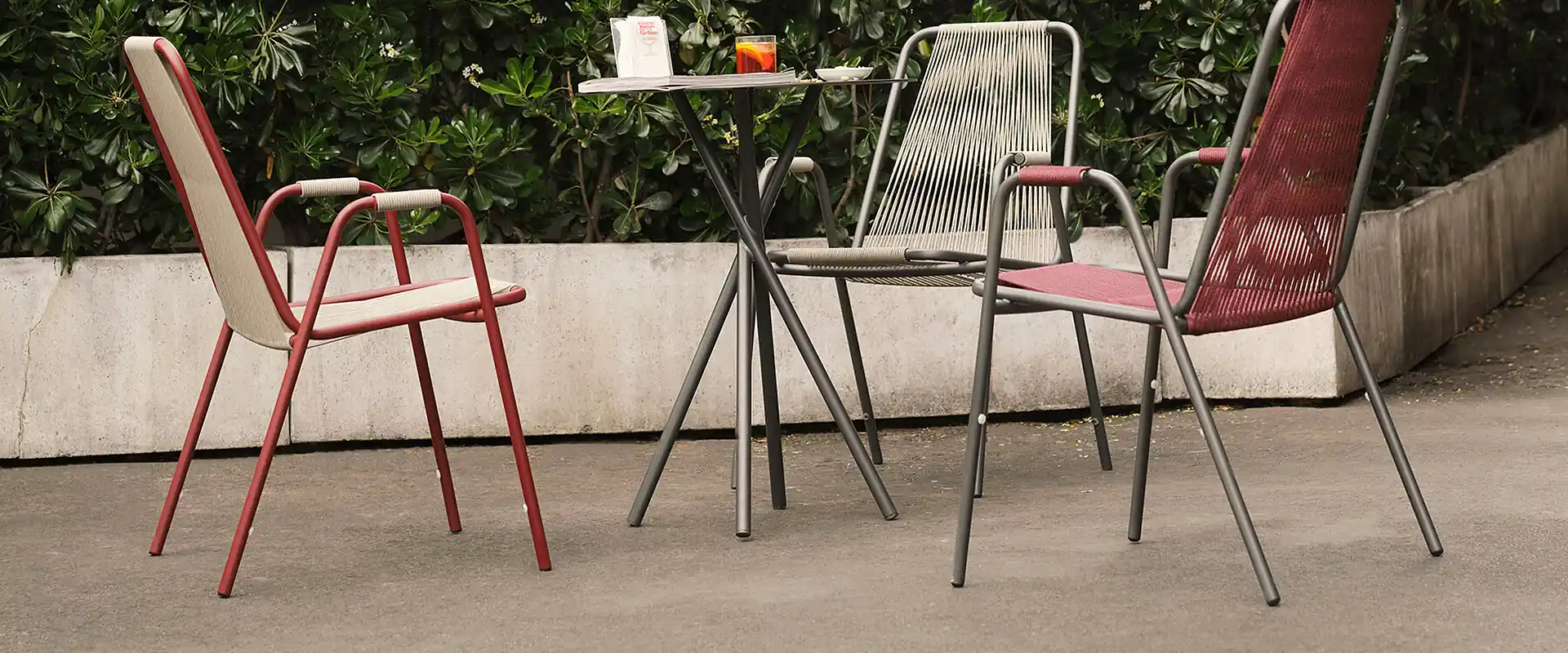 Three Unopiù Coco chairs with woven cord seats in red and beige around a bistro table, in front of a hedge
