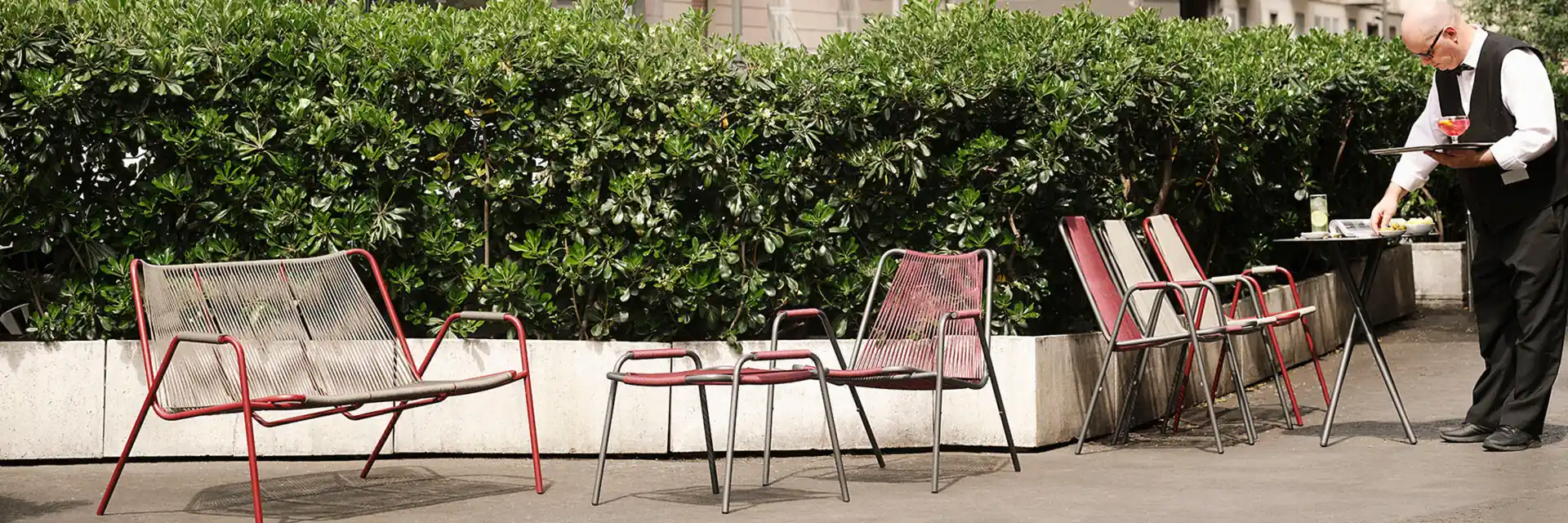 Unopiù Coco woven-cord chairs along a clipped hedge on a city hotel terrace, with a waiter setting a table