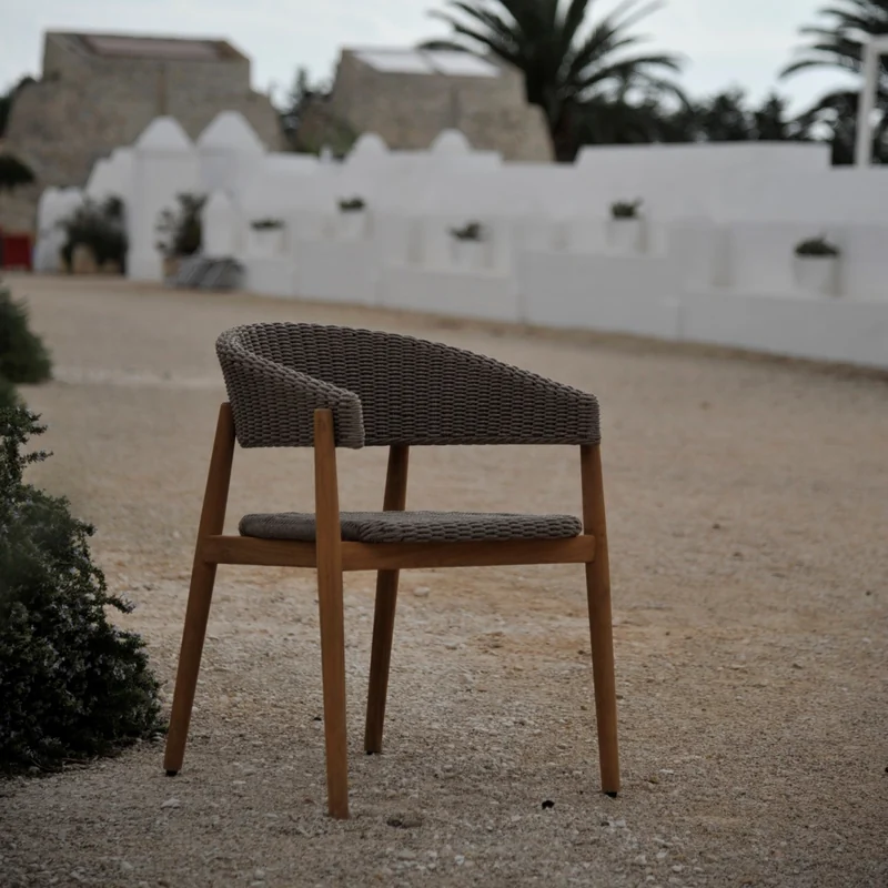 Unopiù Pevero teak armchair with a woven back on a gravel terrace, with white houses and a palm tree behind