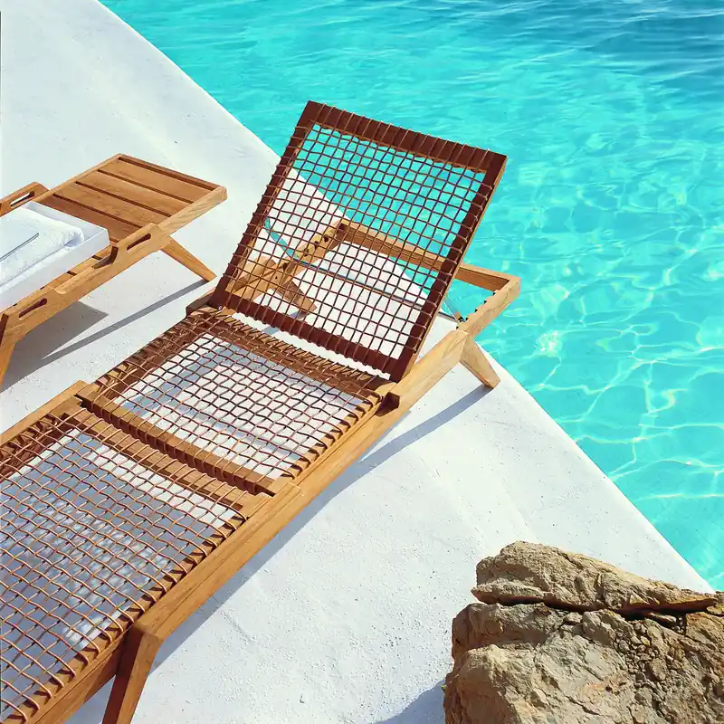 Unopiù Synthesis teak sun lounger with a woven backrest raised, on white stone at the edge of a turquoise pool