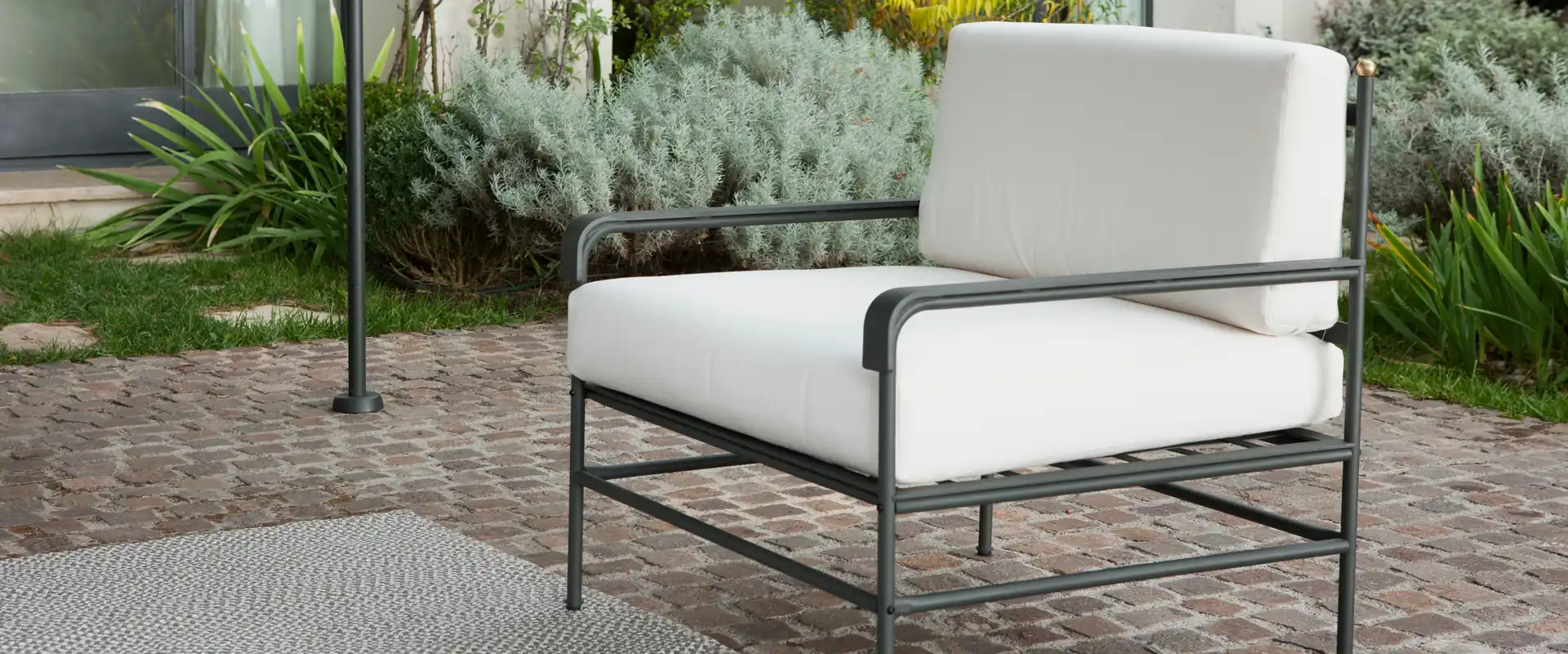 Unopiù Toscana iron armchair with thick white cushions on a stone terrace, with gray-green garden plants behind