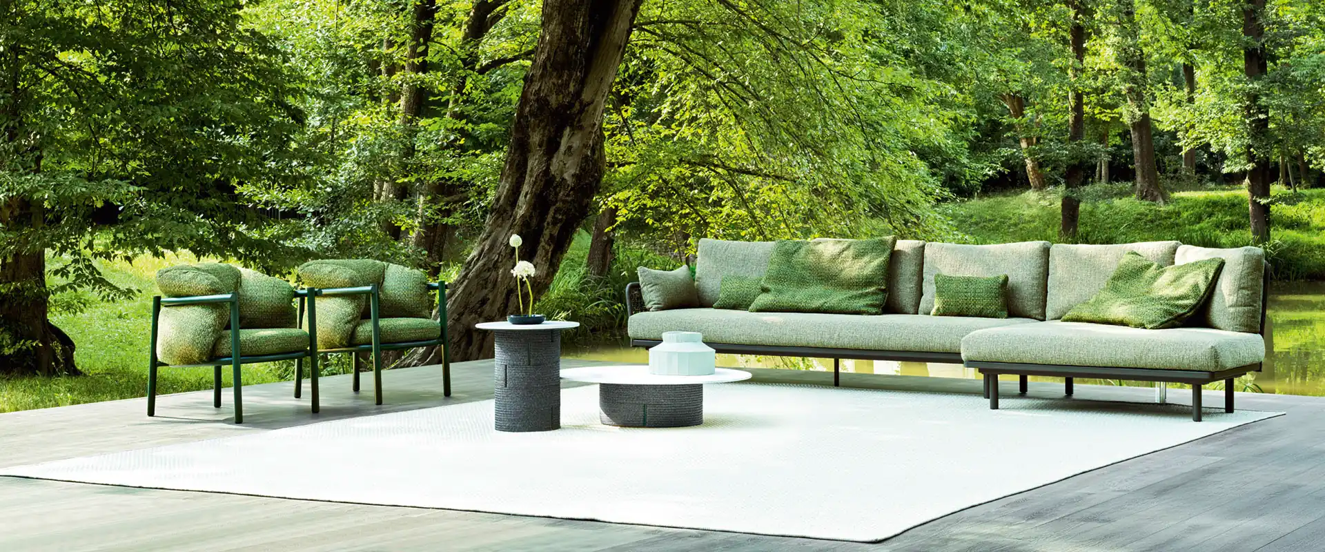 Two Varaschin Allegra armchairs in green beside a sofa and low tables on a timber deck with a white rug, under tall trees by a pond