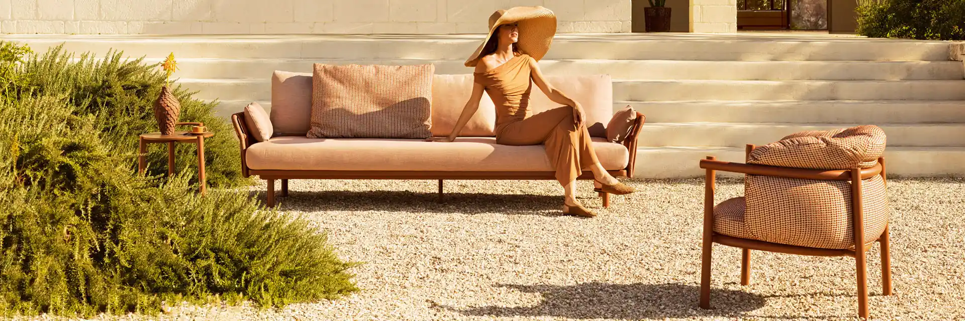 Woman lying on a Varaschin Allegra sofa beside an Allegra armchair, at the foot of the steps of a pale stone house