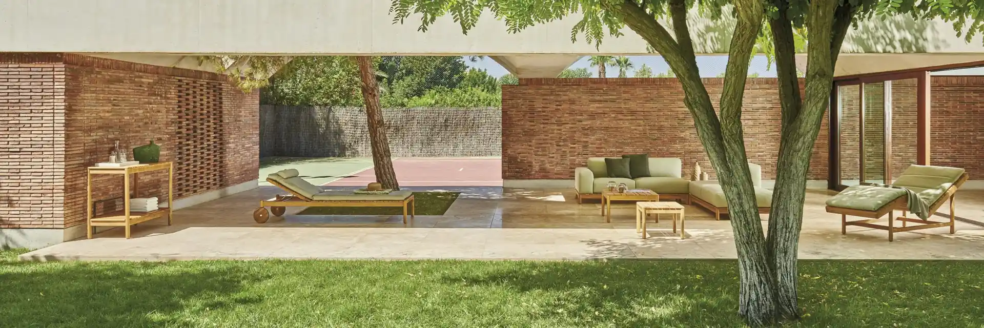Varaschin Barcode sofa and teak sun loungers in an open brick pavilion, with a lawn and a large tree in front