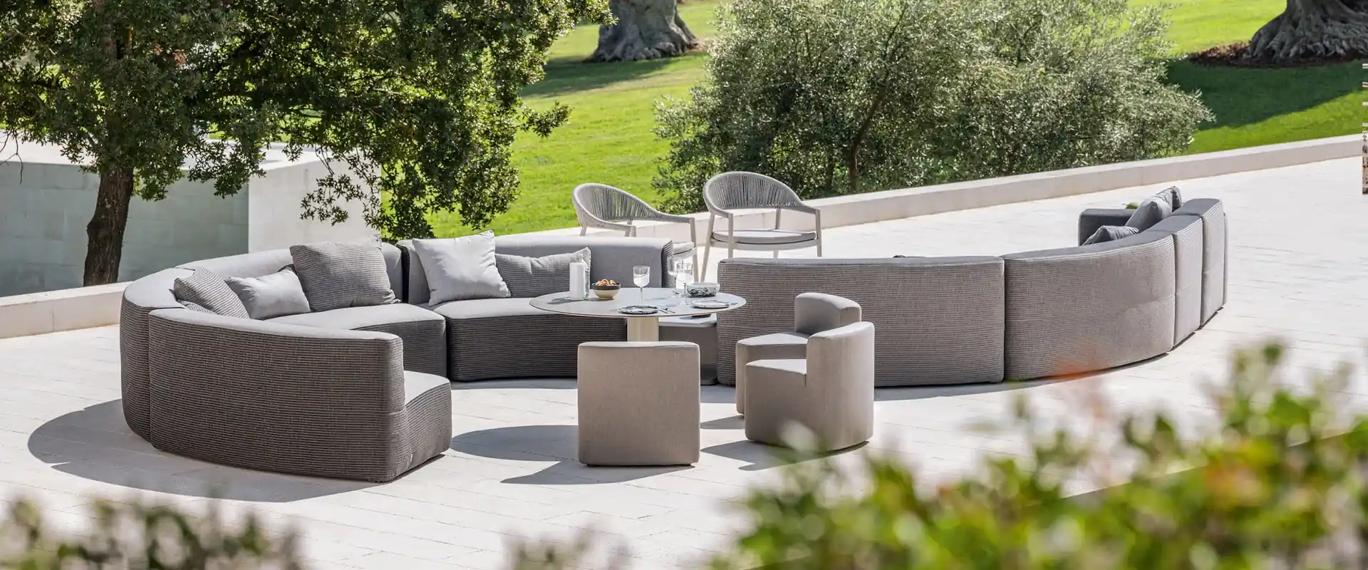 Varaschin Belt curved modular sofa in gray around poufs and low tables on a pale terrace, with olive trees and a lawn behind
