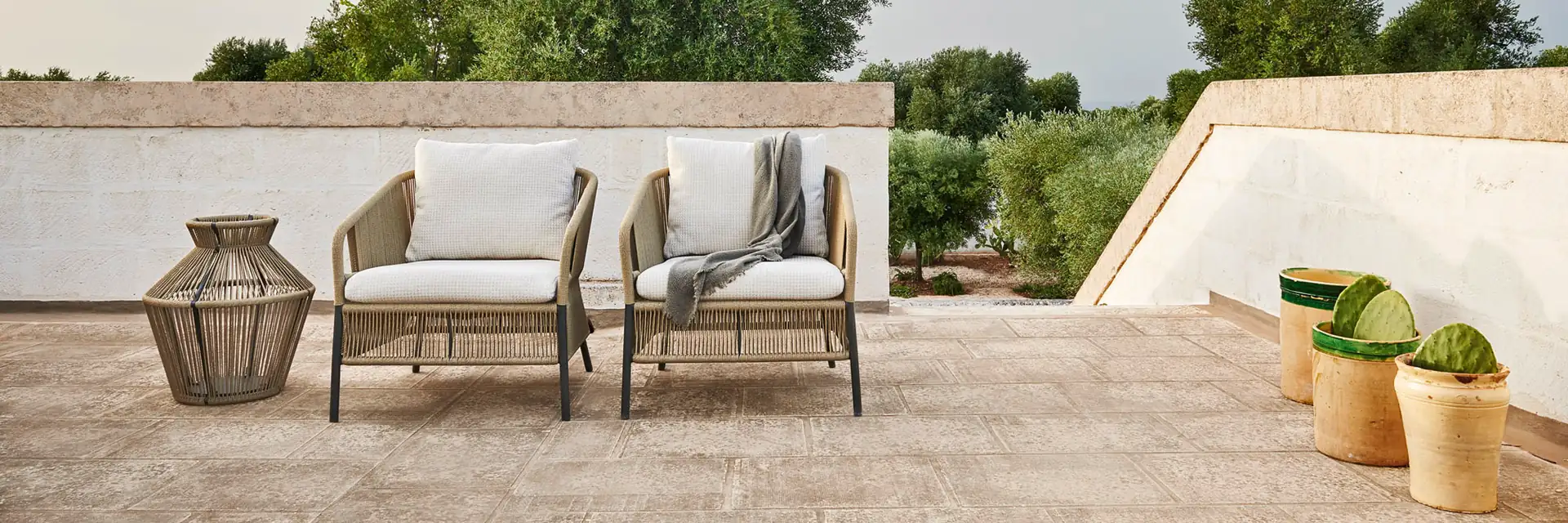 Two Varaschin Cricket armchairs beside a woven lantern on a stone rooftop terrace, with olive trees behind