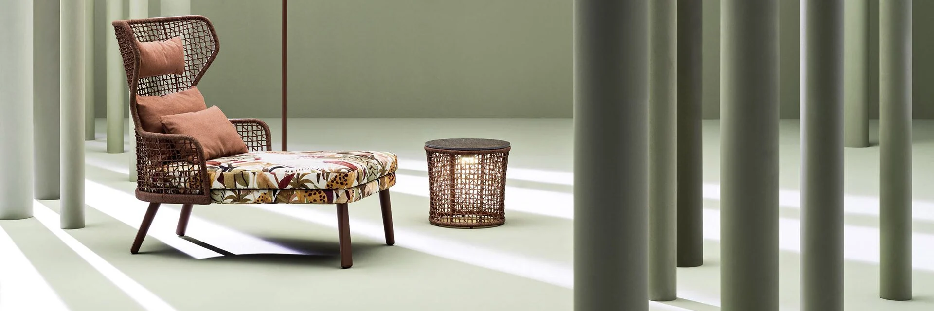Varaschin Emma high-back lounge armchair and footstool with floral cushions beside a woven side table, among sage-green columns