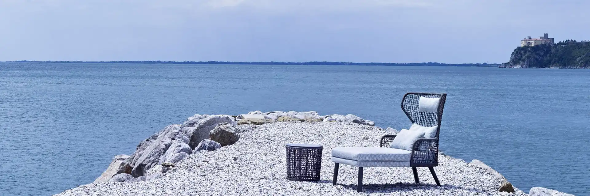 Varaschin Emma high-back lounge chair with a footstool on a pebble point in the sea, with a castle on a distant headland