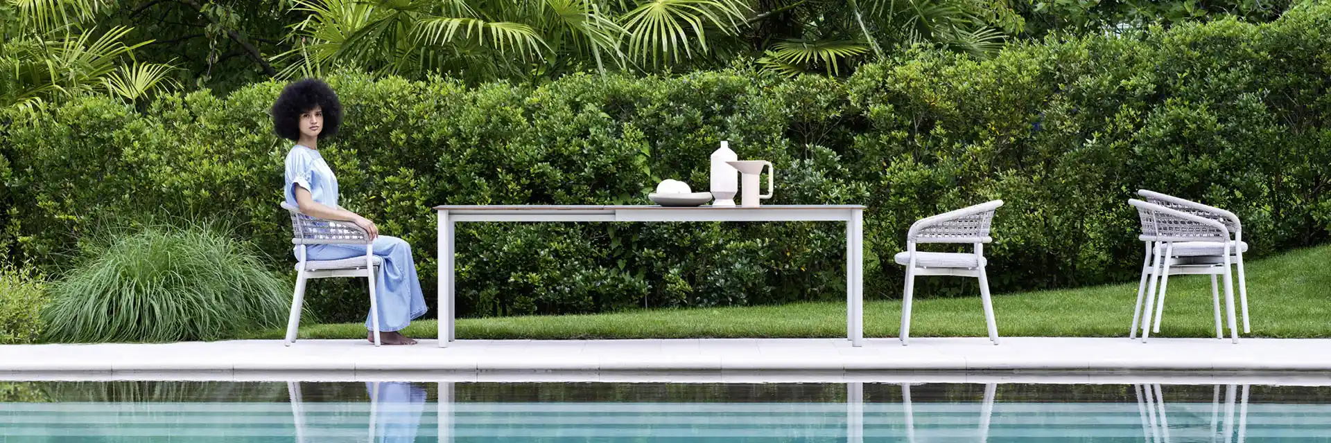 Woman sitting on a Varaschin Plinto dining armchair at a long table beside a pool, with a lush garden behind