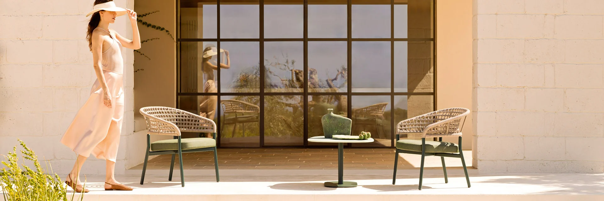 Varaschin Plinto lounge armchairs and a side table on a stone terrace in warm evening light, with a woman walking past a large window