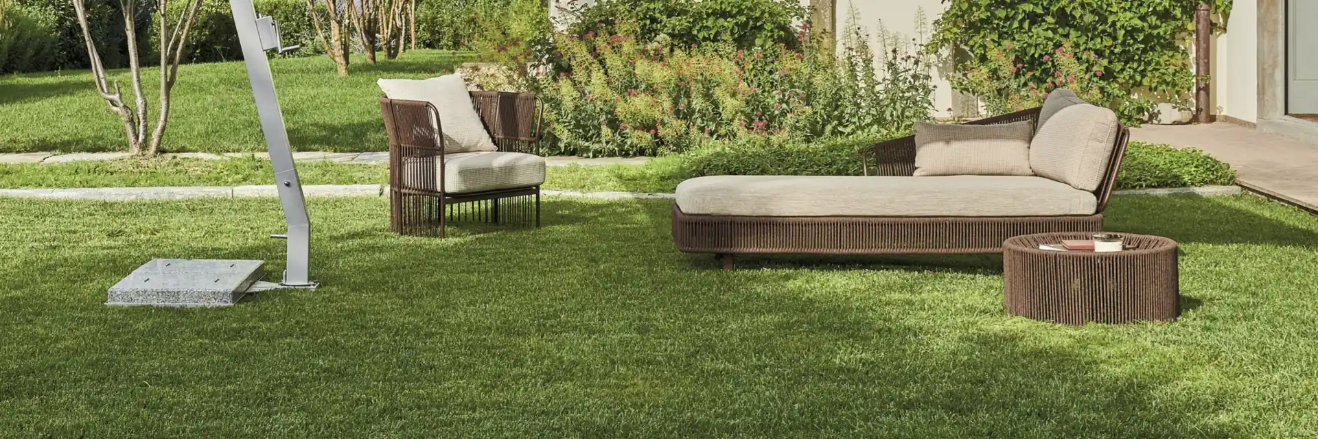 Varaschin Tibidabo woven armchair, chaise longue and round coffee table on the lawn of a pale villa, beside a cantilever parasol