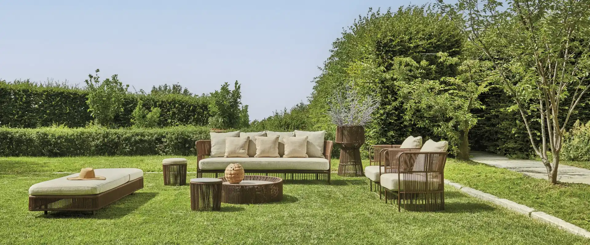 Varaschin Tibidabo woven sofas, armchairs and a sun lounger on a lawn, with a hedge and trees behind