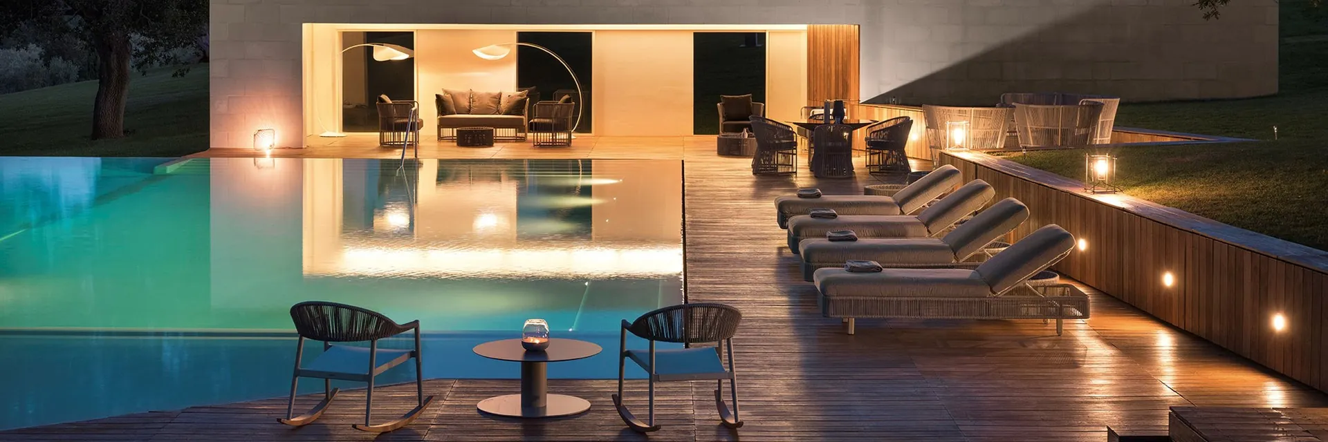 Varaschin Tibidabo armchairs and sun loungers on a deck beside a lit pool house at night